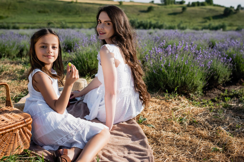 smiling girl holding grape near mother on picnic in flowering field - Foto, imagen