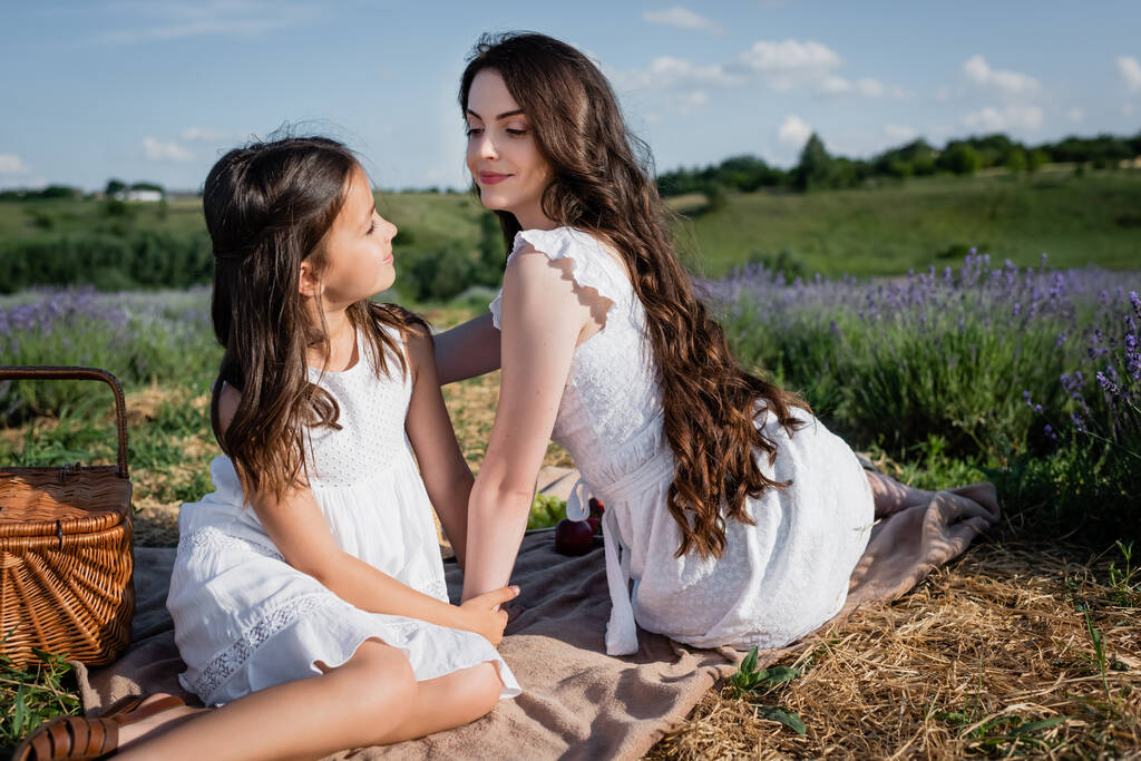 brunette woman and girl smiling at each other while sitting on blanket in countryside - Fotó, kép