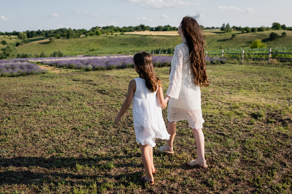 πλήρες μήκος της μητέρας και της κόρης σε λευκά φορέματα με τα πόδια στον τομέα  - Φωτογραφία, εικόνα