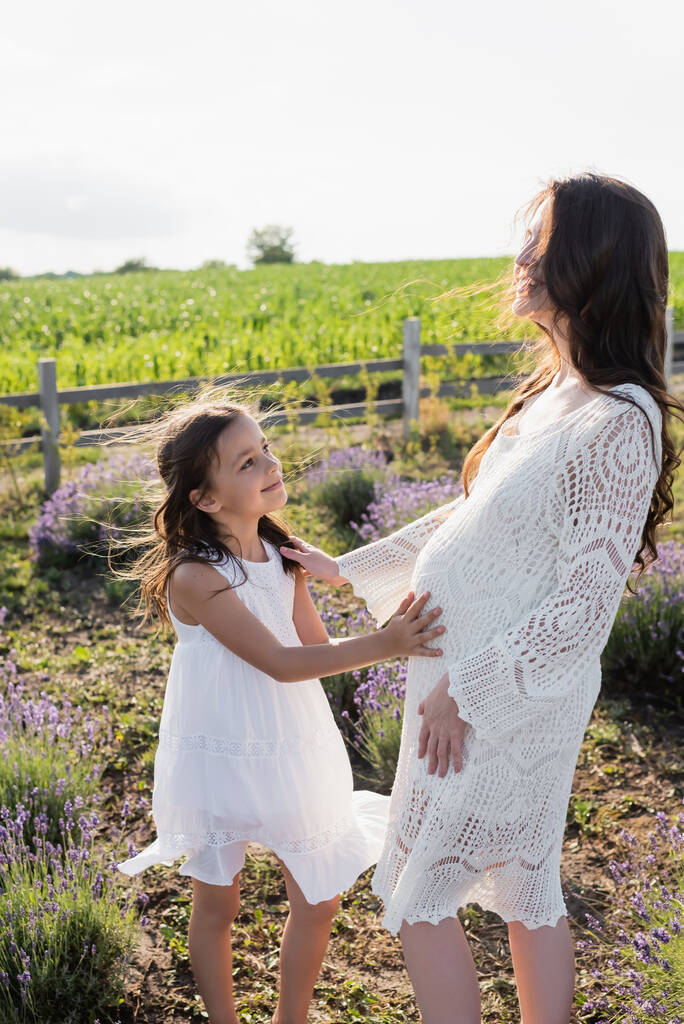 smiling girl touching tummy of pregnant mom in field  - Foto, Imagem