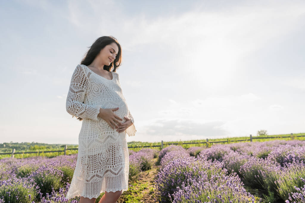 счастливая беременная женщина в платье с открытыми дверями обнимает живот на лавандовом поле - Фото, изображение