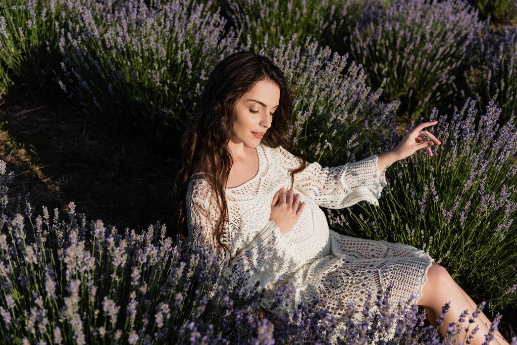 high angle view of pregnant woman with closed eyes touching tummy while sitting in lavender field - Photo, Image