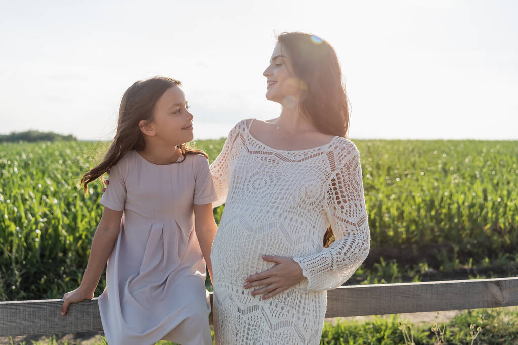 happy pregnant woman and girl in pink dress looking at each other near wooden fence in meadow - Fotoğraf, Görsel
