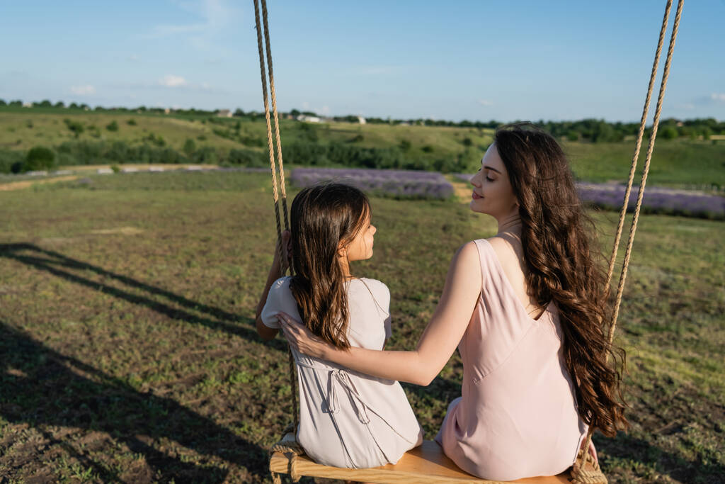 happy brunette woman looking at daughter while riding swing in meadow - Foto, Imagem
