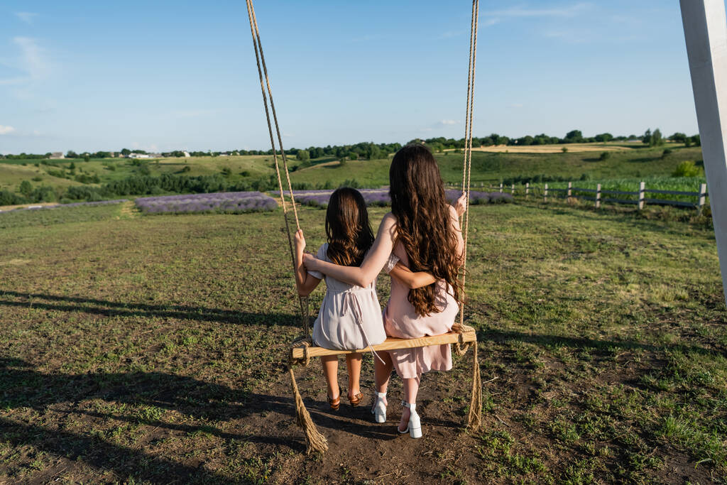 Вид сзади брюнетки и ребенка на качелях для верховой езды в поле - Фото, изображение