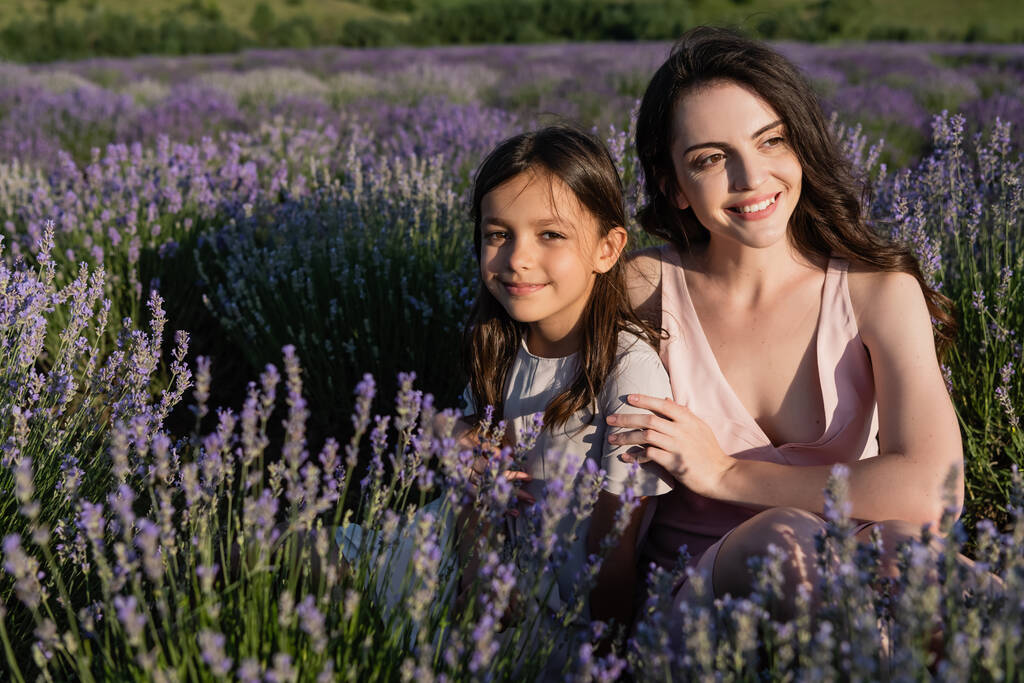 joyful brunette mother and daughter sitting in meadow with flowering lavender - Valokuva, kuva