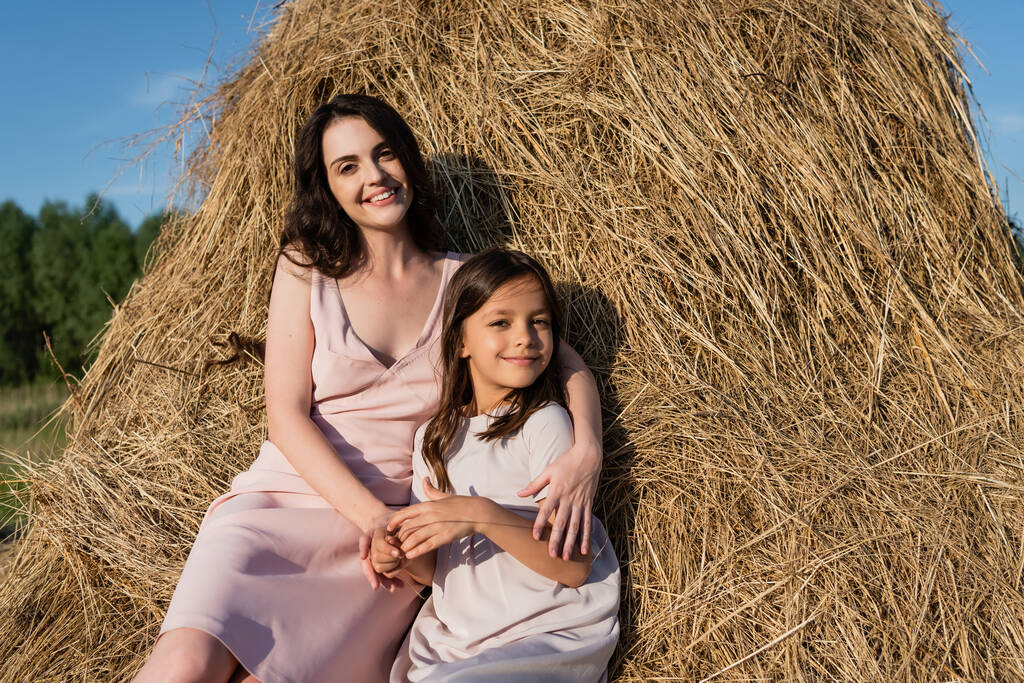 woman and daughter in pink dresses looking at camera while sitting on haystack - Foto, afbeelding