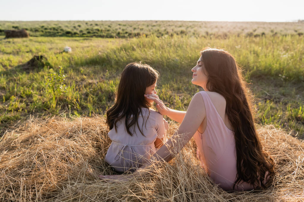 brunette woman touching face of daughter while sitting on haystack in field - Photo, image