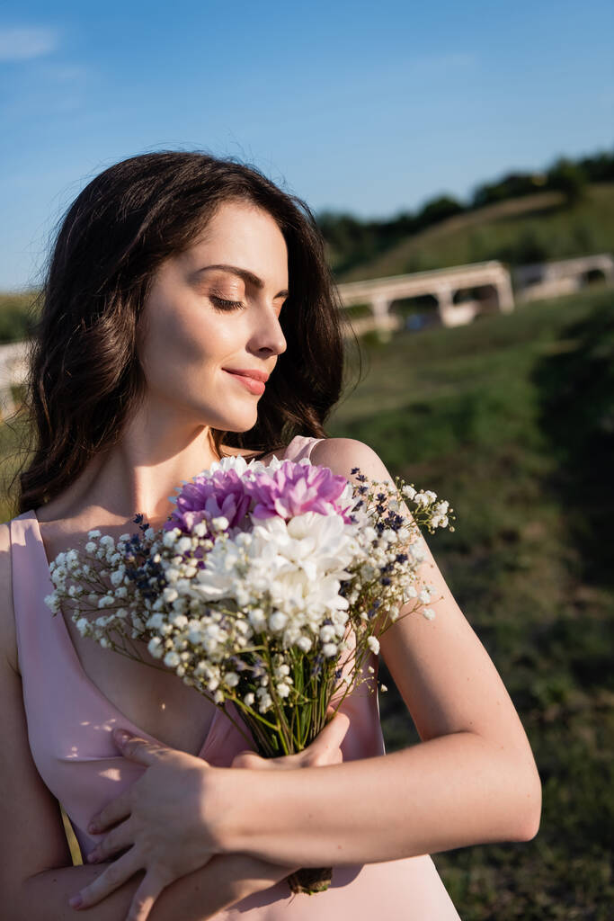 брюнетка женщина с букет летних цветов улыбается на открытом воздухе - Фото, изображение