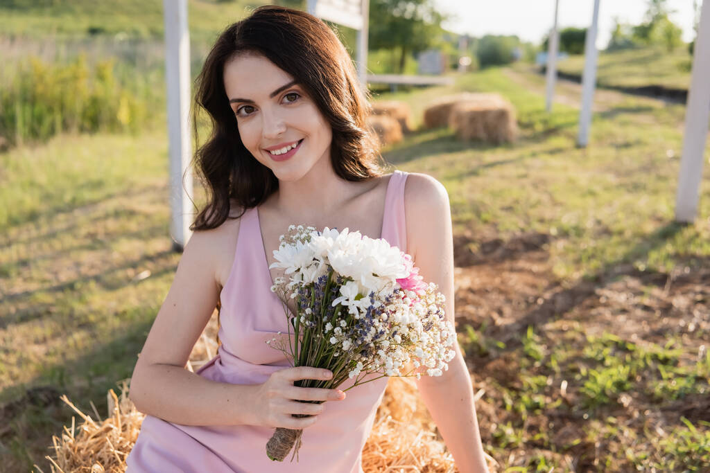 cheerful brunette woman with bouquet smiling at camera in countryside - Fotografie, Obrázek