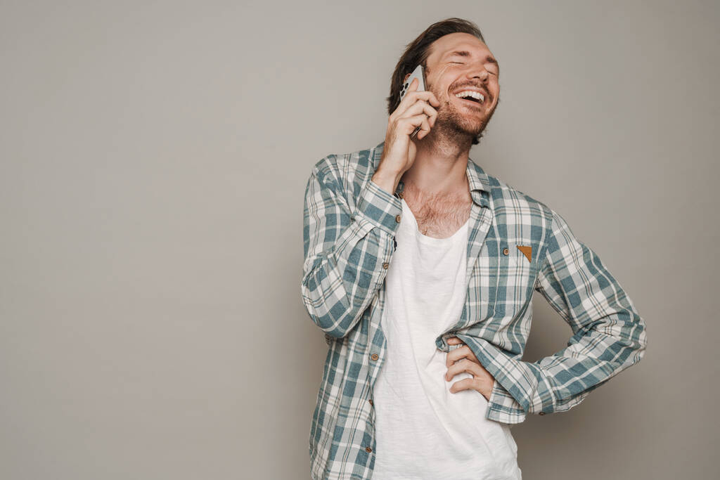 Bristle man dressed in shirt laughing while talking on cellphone isolated over grey background - Foto, Bild