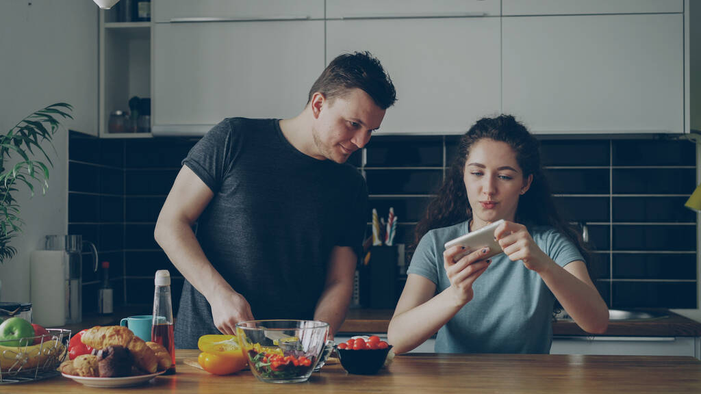 Couple attrayant dans la cuisine à la maison. Fille bouclée jouer jeu vidéo sur smartphone tandis que son copain cuisine petit déjeuner
 - Photo, image