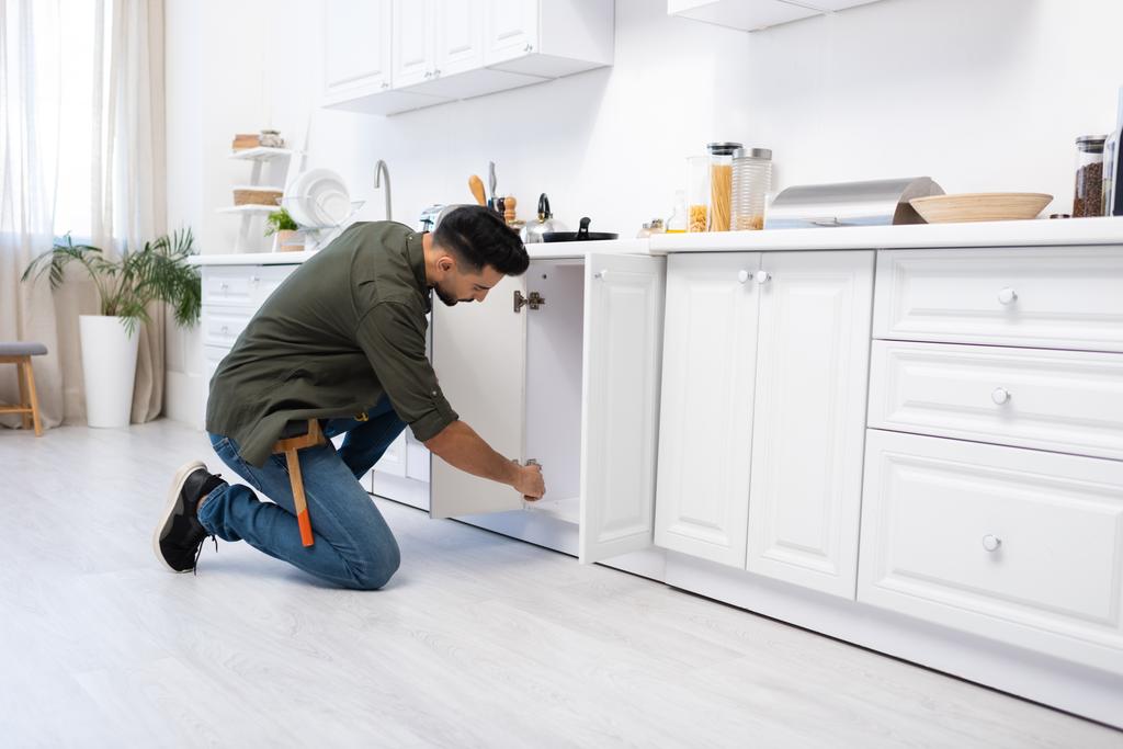 Πλευρική άποψη του muslim άνθρωπος με ζώνη εργαλείων στερέωση ντουλάπι κουζίνα  - Φωτογραφία, εικόνα