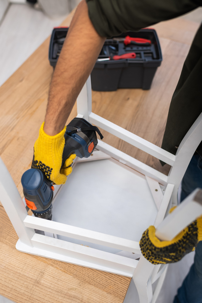 Обрезанный вид человека в перчатках ремонтный стул с электрической отверткой возле размытого ящика инструментов дома  - Фото, изображение