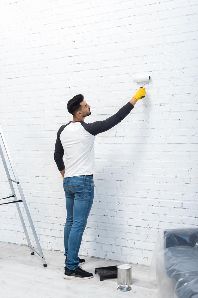 Side view of young muslim man coloring wall near dye and ladder at home  - Foto, imagen
