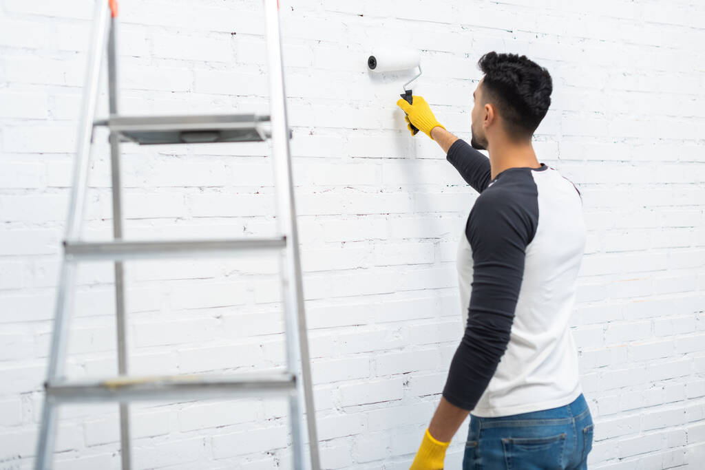Arabian man in gloves coloring wall near ladder at home  - Photo, Image