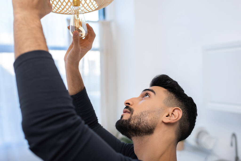 Side view of arabian man changing lightbulb in chandelier in kitchen  - Fotografie, Obrázek
