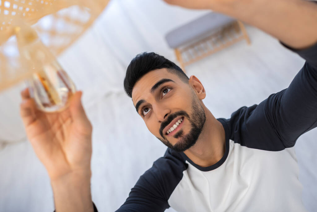 High angle view of cheerful muslim man changing lightbulb in blurred chandelier at home - Foto, afbeelding