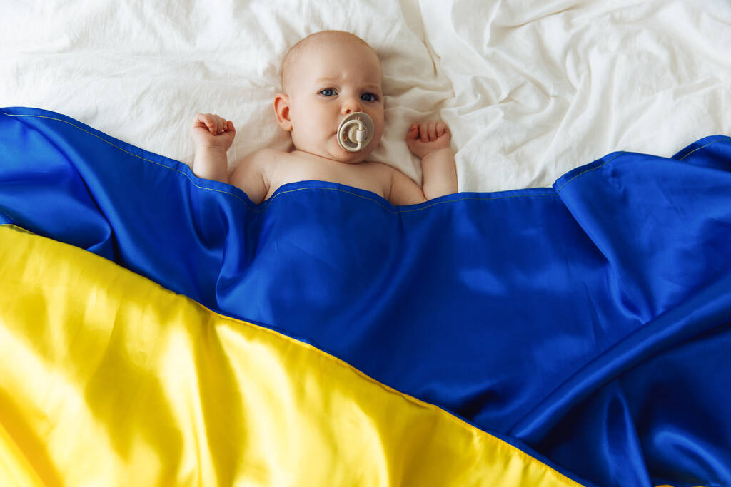 Portrait of a baby wrapped in the national blue and yellow flag of Ukraine, lying on the bed. Baby with Ukraine flag heart. photo with place for text. Children of war. The child is scared. Hope for the world. fear and expectation. children's tears. S - Fotó, kép