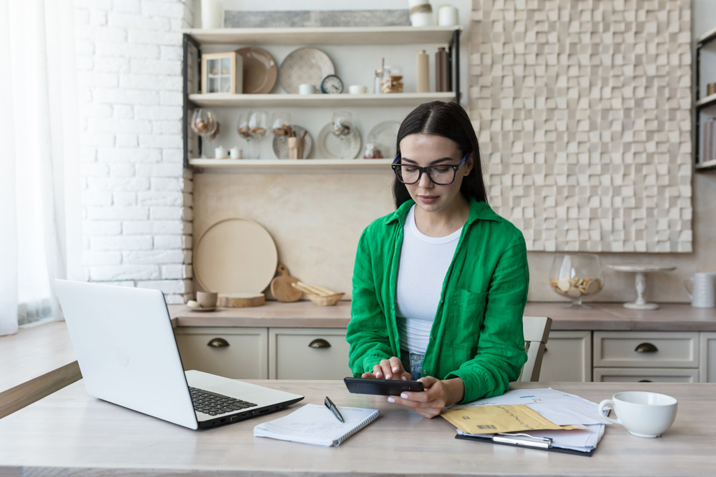 Serious and focused businesswoman freelancer on paper work at home in kitchen, brunette in glasses and green in kitchen, paying and calculating bills - 写真・画像
