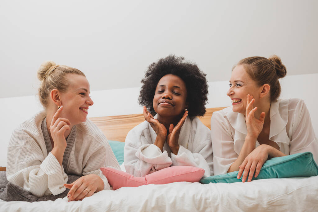 Οι νεαρές γυναίκες είναι ξαπλωμένες με μπουρνούζια στο κρεβάτι και επικοινωνούν. Οι χαρούμενες φίλες χαλαρώνουν στο ξενοδοχείο σπα. - Φωτογραφία, εικόνα
