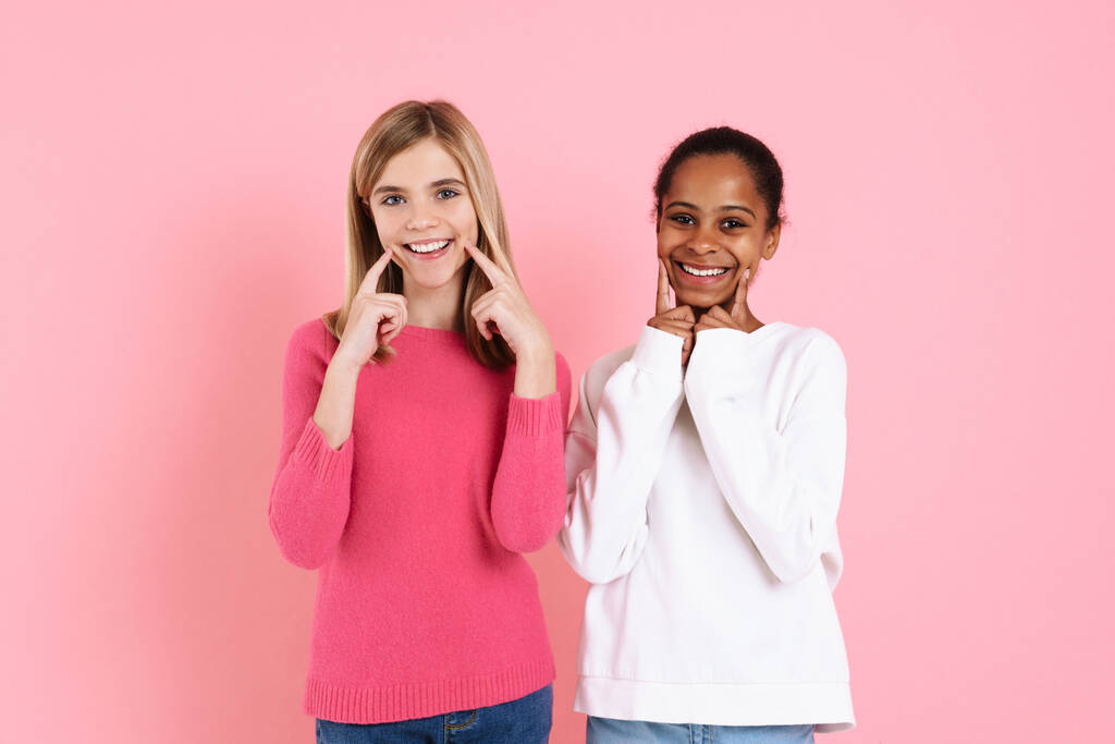 Multiracial two girls wearing sweaters pointing fingers at their smiles isolated over pink wall - Foto, afbeelding