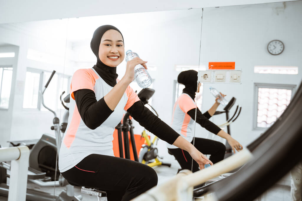 красива мусульманка п'є пляшку води, використовуючи статичний велосипед у спортзалі сама
 - Фото, зображення