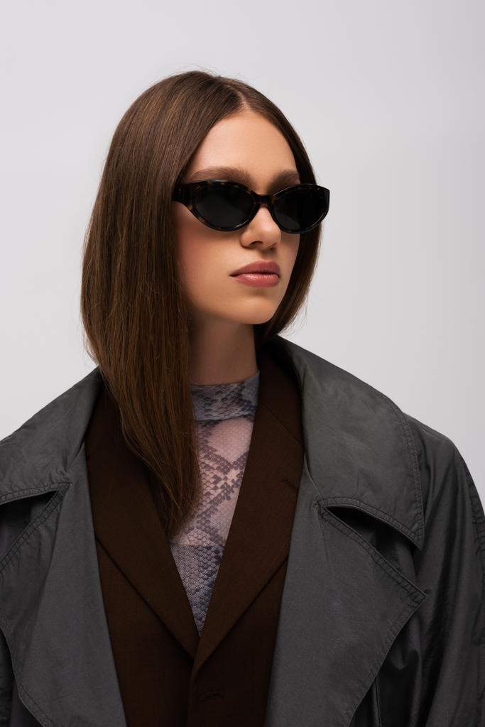 μελαχρινή εφηβική μοντέλο σε μοντέρνα γυαλιά ηλίου και μοντέρνα καμπαρντίνα απομονώνονται σε γκρι - Φωτογραφία, εικόνα