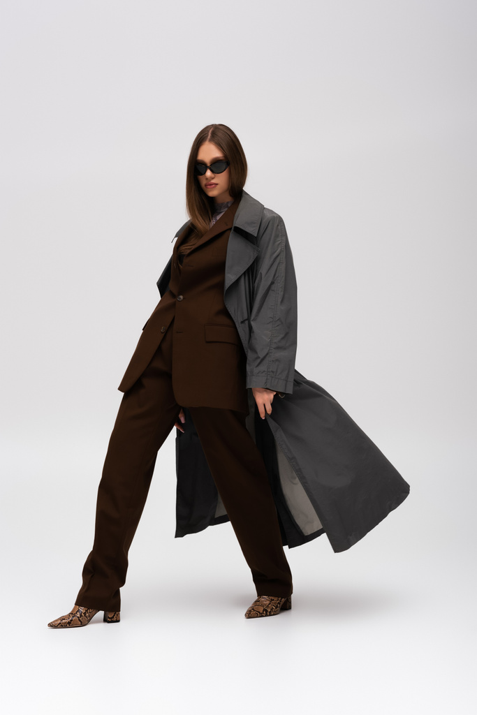 full length of stylish teenage model in sunglasses and trendy trench coat walking on grey - 写真・画像
