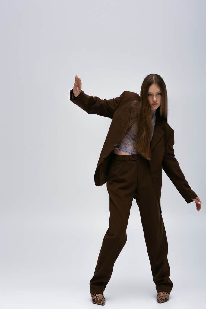 full length of stylish teenage model in brown high-quality suit gesturing on grey - 写真・画像