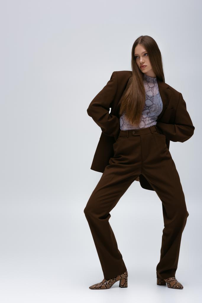 full length of trendy teenage model in brown high-quality suit posing on grey - Fotó, kép