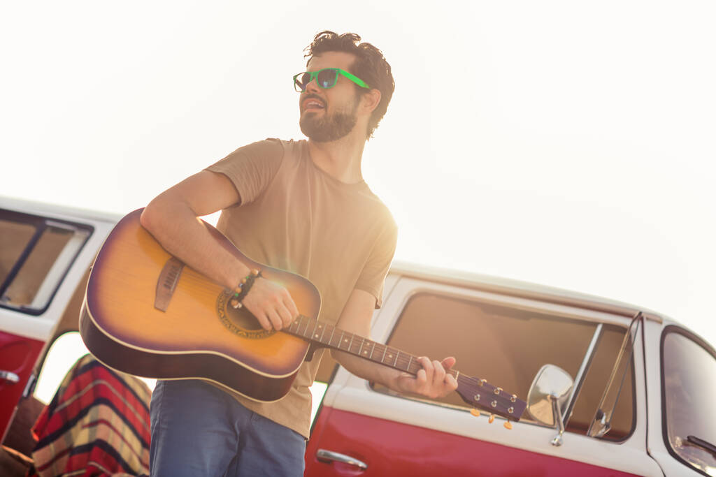 Фото смішного красивого безтурботного хіпі хлопця посміхається грати на гітарі верхи ретро фургон за межами приморського пляжу
. - Фото, зображення