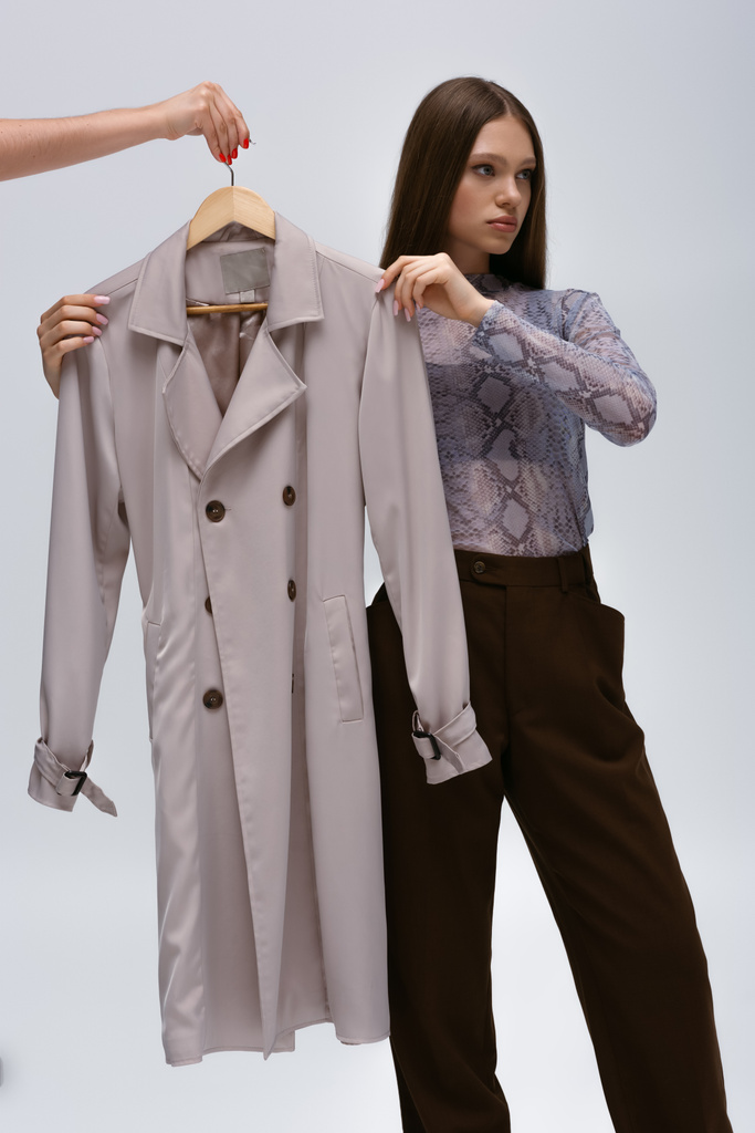 teenage model and woman hugging stylish trench coat on hanger isolated on grey - Photo, Image