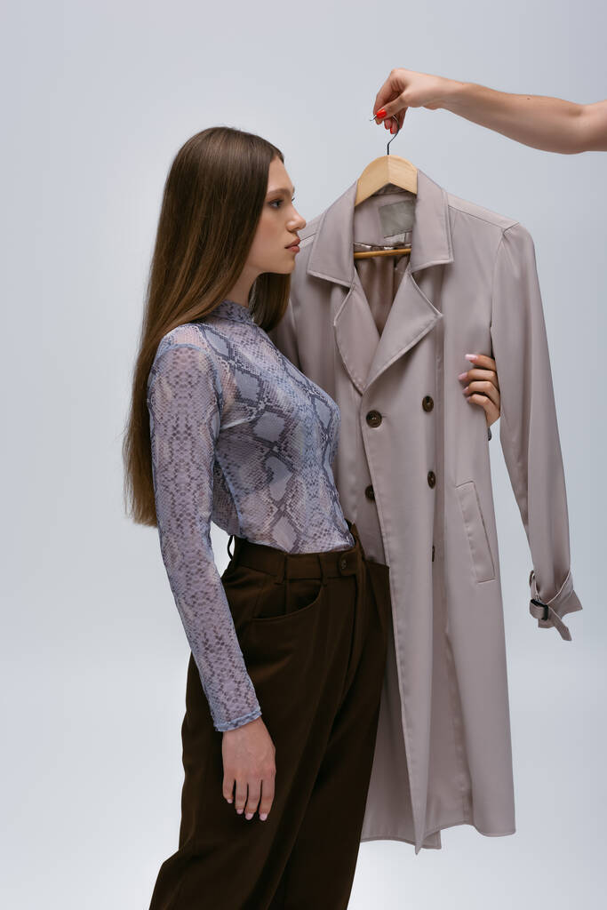 side view of teenage model and woman holding recyclable trench coat on hanger isolated on grey - Photo, Image
