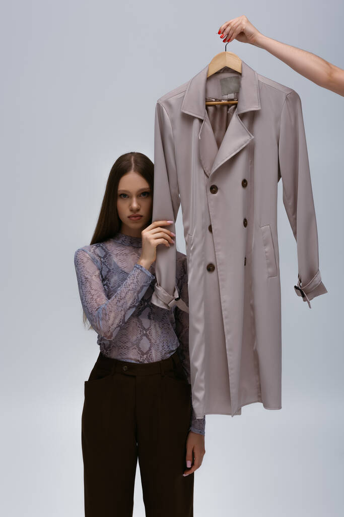 підліткові моделі торкаються рукава перероблюваного траншеї пальто, а жінка тримає вішалку ізольовано на сірому
 - Фото, зображення