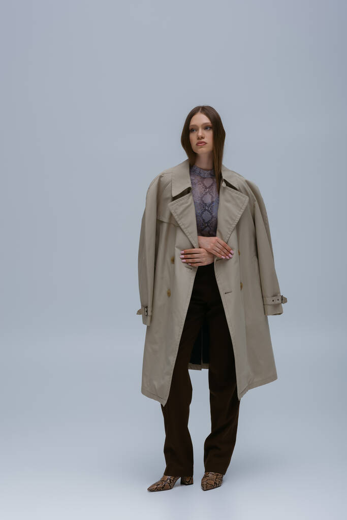 full length of teenage model posing in stylish trench coat on grey  - Foto, immagini