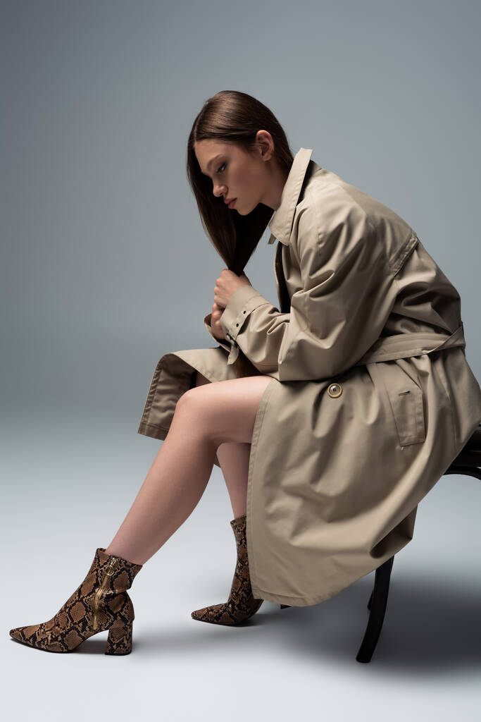full length of model in stylish trench coat sitting on chair and posing on grey - 写真・画像