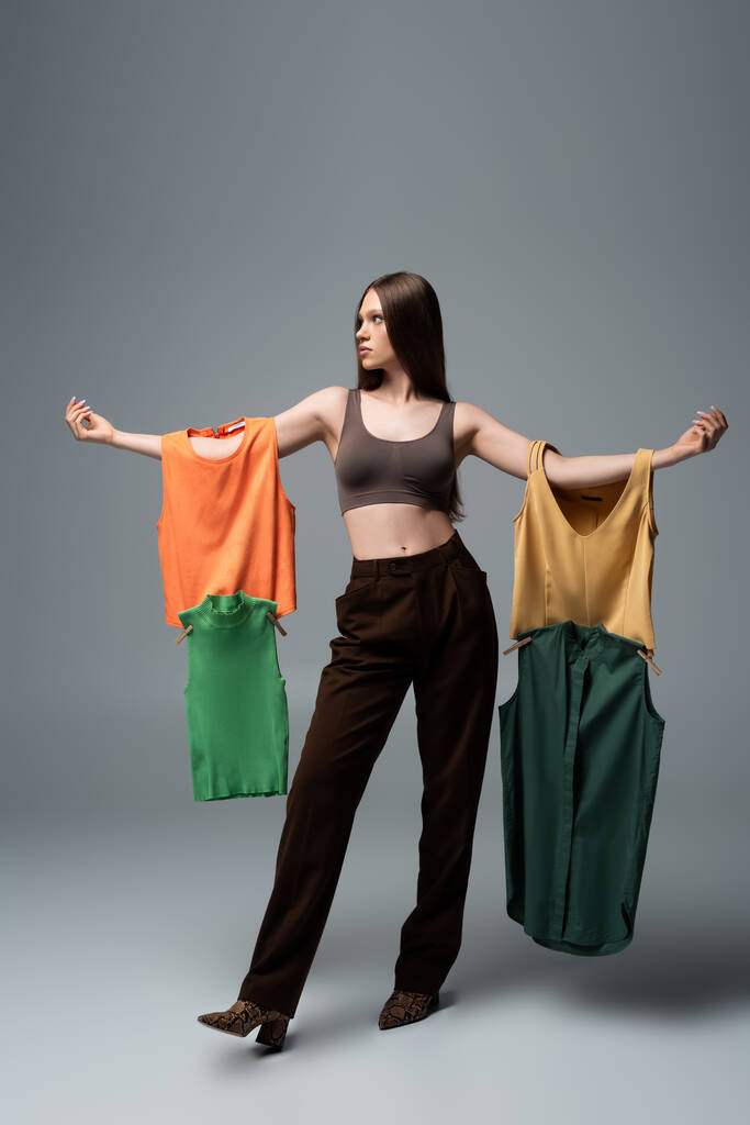 full length of pretty model in crop top and trousers standing with outstretched hands while holding clothing on grey  - Photo, Image