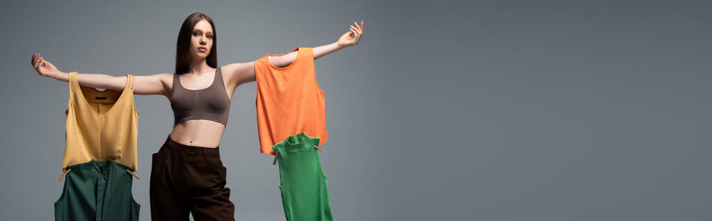 красивая модель в кукурузном топе и брюках, стоящих с протянутыми руками и держащих одежду на сером, баннер - Фото, изображение