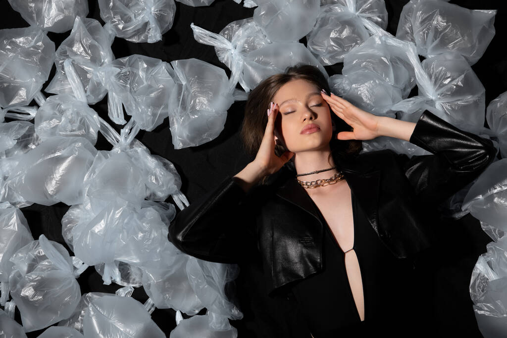top view of brunette model with closed eyes lying around plastic bags on black  - Fotoğraf, Görsel