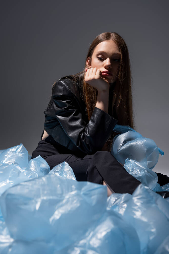 грустная модель в стильном черном костюме, позирующая вокруг синих пластиковых пакетов на сером - Фото, изображение