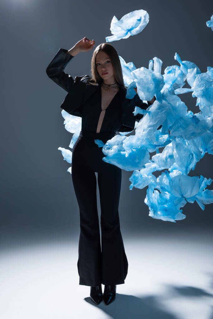 πλήρες μήκος του μοντέλου μελαχρινή σε κομψό κοστούμι ποζάρουν κοντά σε ιπτάμενες μπλε πλαστικές σακούλες σε σκούρο γκρι - Φωτογραφία, εικόνα