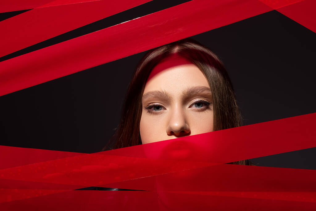 teenage model looking at camera through red strips from adhesive tape isolated on black, recycle fashion concept - 写真・画像