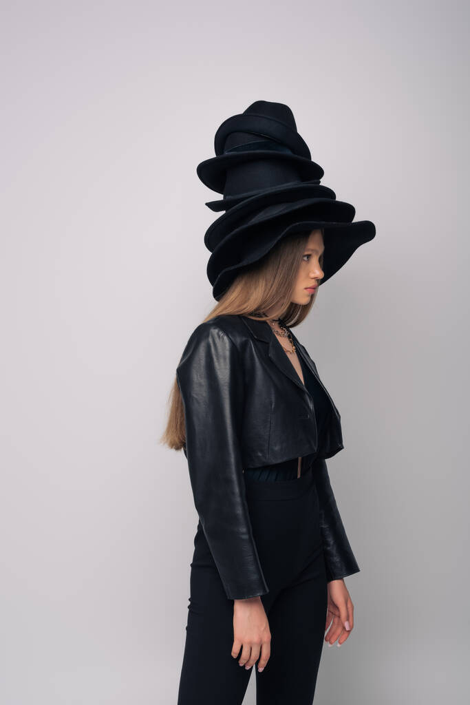 πλαϊνή όψη του μοντέλου μελαχρινή σε δερμάτινο μπουφάν και διάφορα μαύρα καπέλα στο κεφάλι απομονώνονται σε γκρι - Φωτογραφία, εικόνα