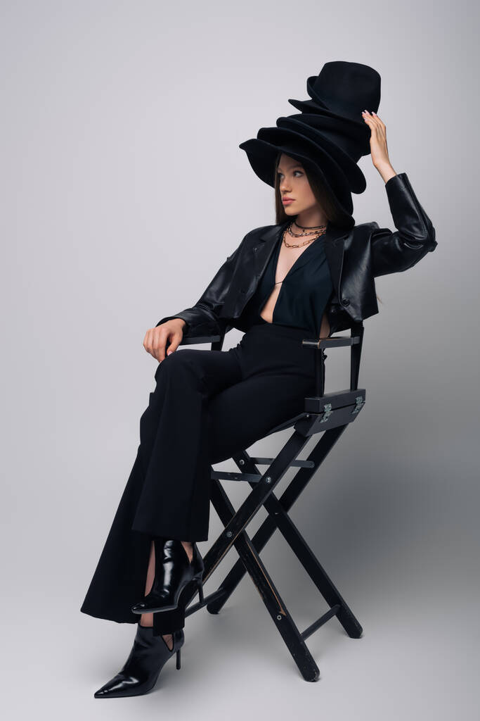 μελαχρινή μοντέλο σε δερμάτινο μπουφάν και διάφορα μαύρα καπέλα κάθεται σε καρέκλα σε γκρι - Φωτογραφία, εικόνα