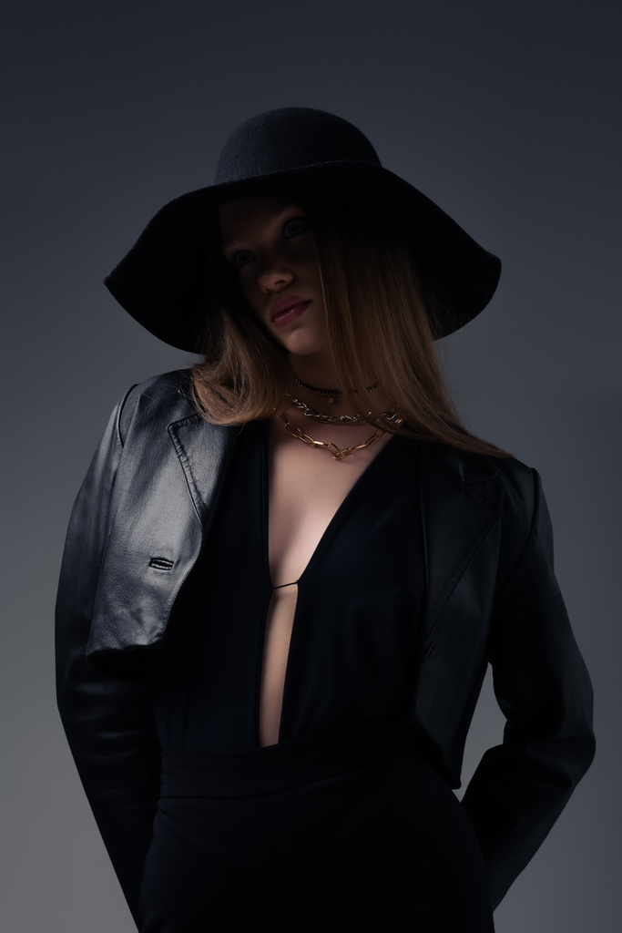 stylish model in floppy hat and black leather jacket isolated on grey - Zdjęcie, obraz