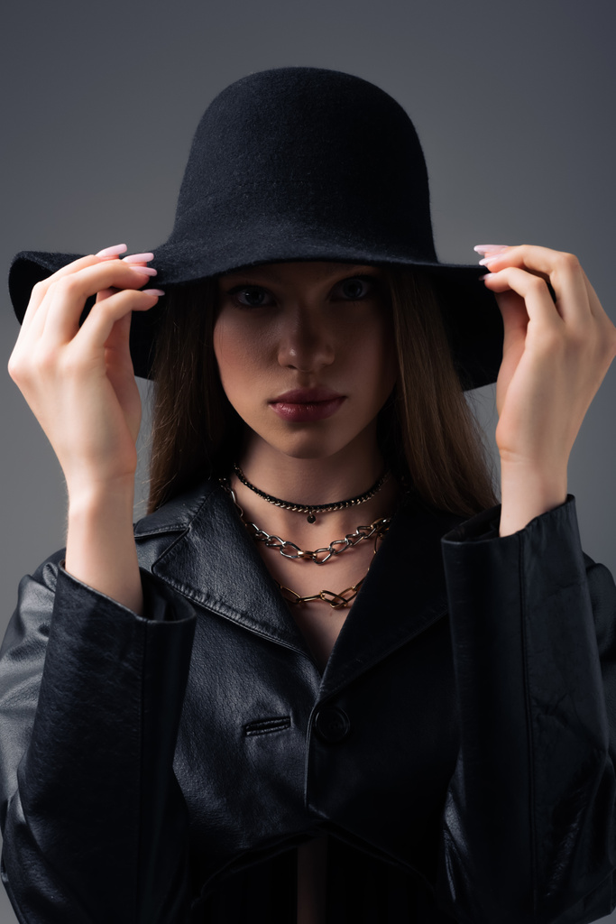 модель подростка в черной кожаной куртке, регулирующей гибкую шляпу, изолированную на сером - Фото, изображение