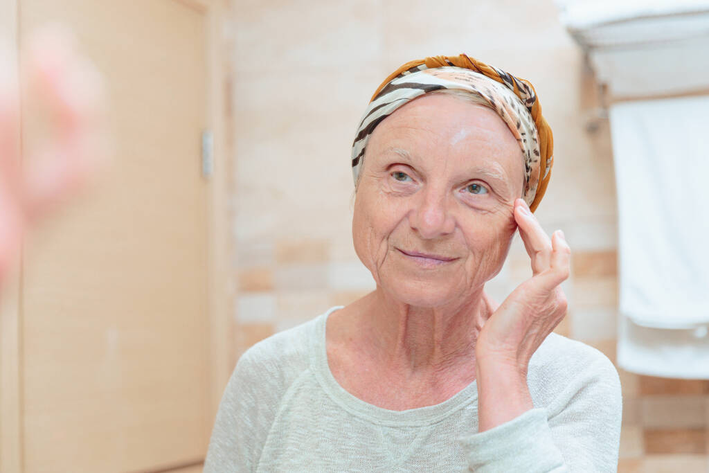 Mature woman taking care of her face. Senior woman standing at the mirror in the bathroom and applying cream or mask on her face - Fotoğraf, Görsel