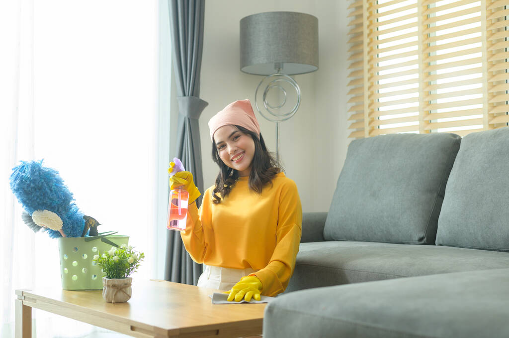 Молода щаслива жінка в жовтих рукавичках і стирає стіл у вітальні
. - Фото, зображення