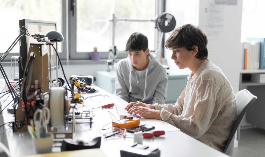 Студенти, які сидять за столом в лабораторії і вивчають 3D друк разом, один друкує, а інший допомагає йому
 - Фото, зображення